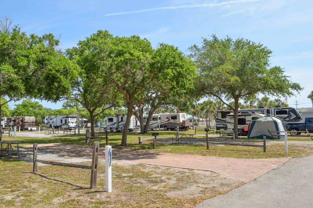 Rows of RVs and a tent are parked under trees at a campground, with picnic tables and paved walkways visible.