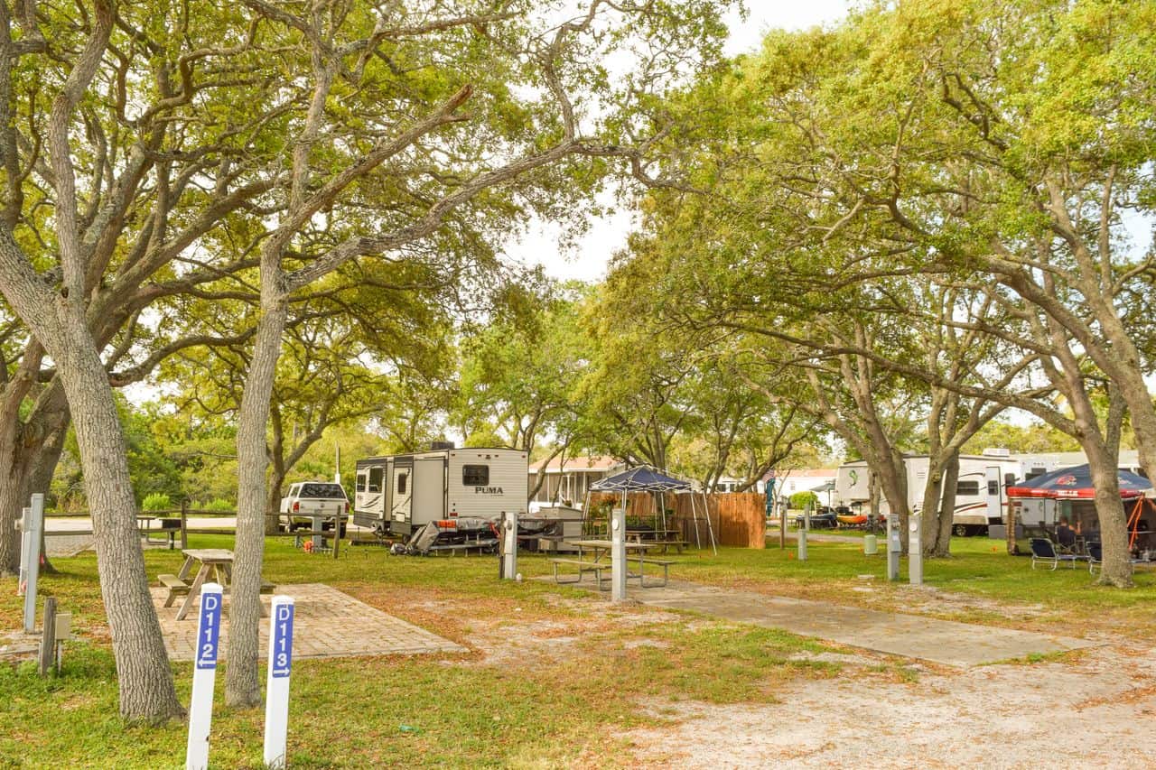 A campground with RVs, trees providing shade, picnic tables, and numbered site markers, under a partly cloudy sky.