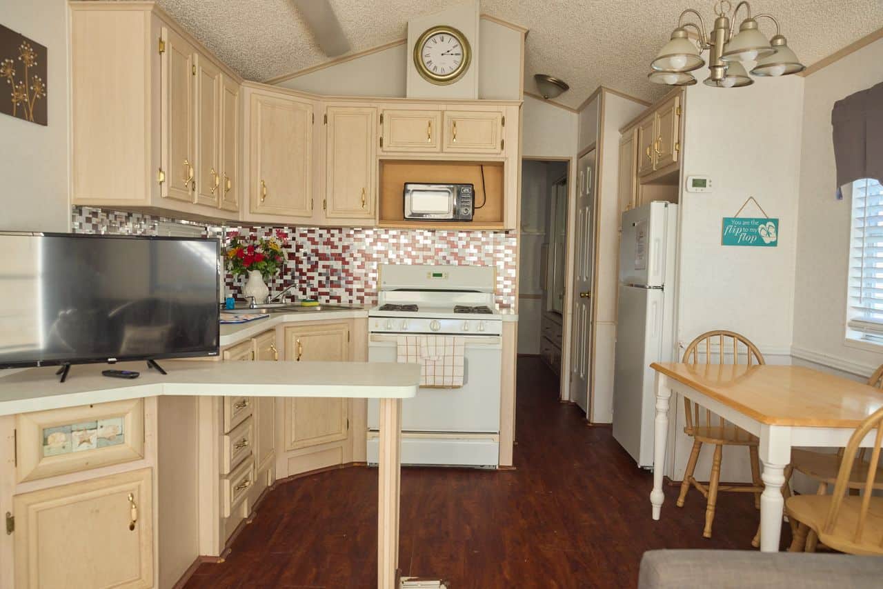 Compact kitchen and dining area with light wood cabinets, stove, microwave, refrigerator, TV, dining table, and wall clock, featuring a mosaic backsplash and hardwood floor.