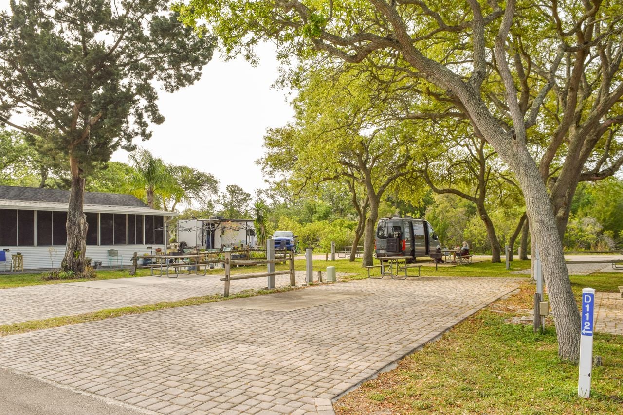 Paved RV campsite with picnic tables and trees, a black camper van parked in the background, and a white building to the left. Site marker labeled D112 in the foreground.