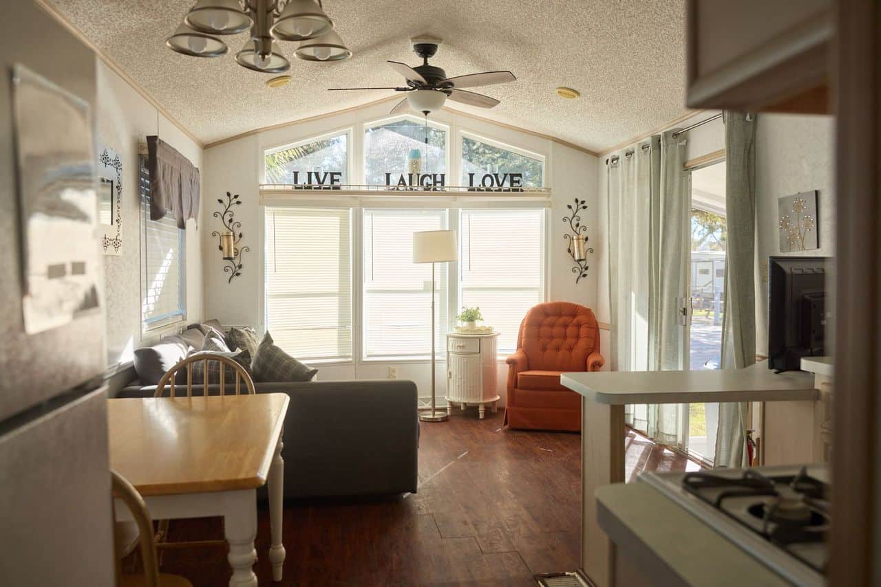 A sunlit living room with a dining table, orange armchair, couch, and large windows. Wall decor reads "Live Laugh Love." Kitchen area and stove are partially visible.
