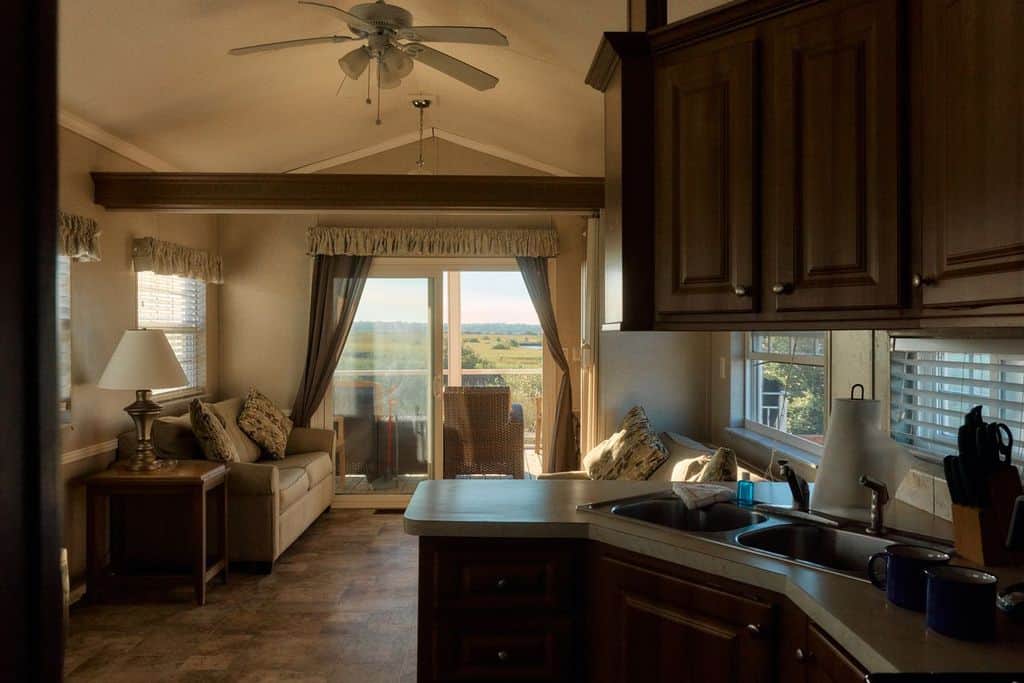 A small, sunlit living room with a sofa, lamp, and kitchen area; glass doors open to a balcony with a view of fields and distant water.