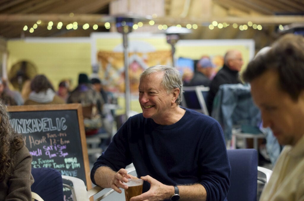 A man sits at a table in a busy cafe, smiling and holding a cup, with a chalkboard menu and string lights visible in the background.