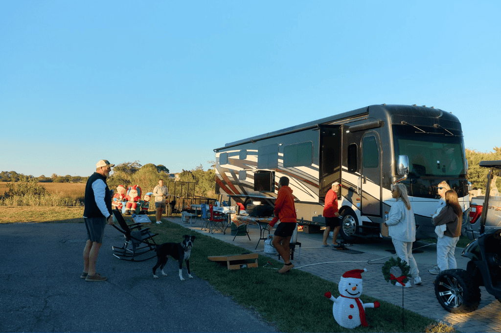 A group of people and a dog gather near a parked recreational vehicle, with Christmas-themed lawn decorations nearby.