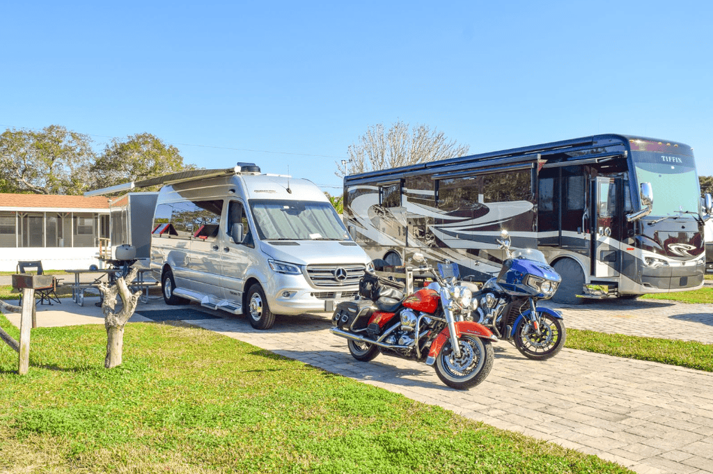 A silver camper van and a large Tiffin motorhome parked on a paved lot, with an orange and a blue motorcycle parked in front of them.