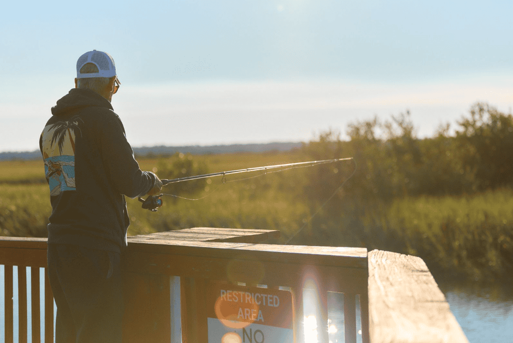 A person wearing a hat and hoodie stands on a wooden pier, fishing in a marshland near a “Restricted Area” sign.