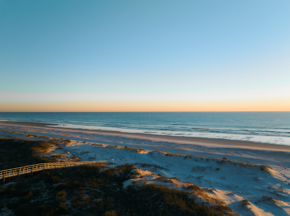 Aerial view of a wooden boardwalk leading through sand dunes to a beach at sunrise.